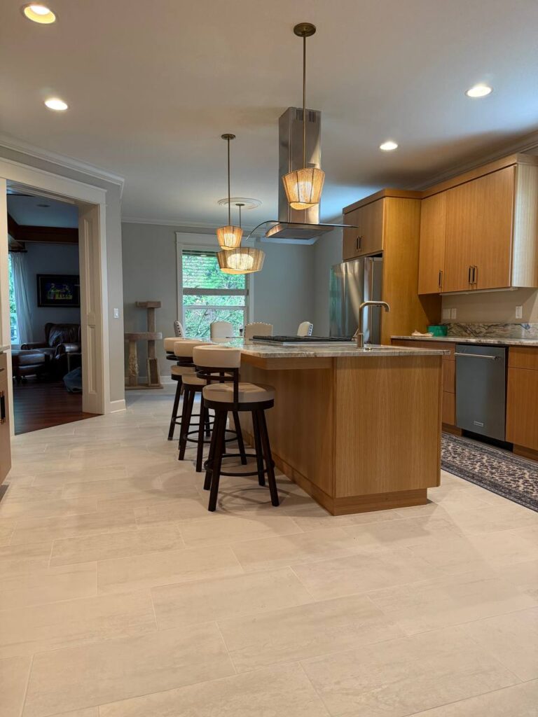 4983430754209565634 Modern kitchen with light wood cabinets, an island with three bar stools, pendant lights, stainless steel appliances, and a window with a view of greenery. A cat tree is visible near an adjoining living room.