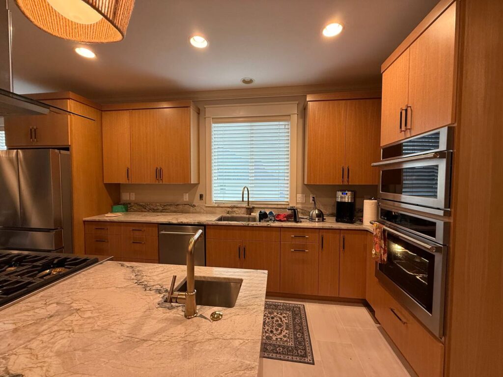 Modern kitchen with wooden cabinets, marble countertops, stainless steel refrigerator, built-in ovens, gas stove, and a sink under a window with white blinds—an ideal result of a quality kitchen renovation in Vancouver WA.