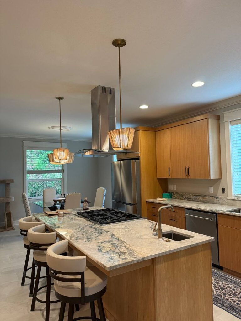 Modern kitchen with light wood cabinets, marble countertops, a central island with bar stools and a gas cooktop, stainless steel appliances, pendant lights, and a dining table with upholstered chairs near large windows.